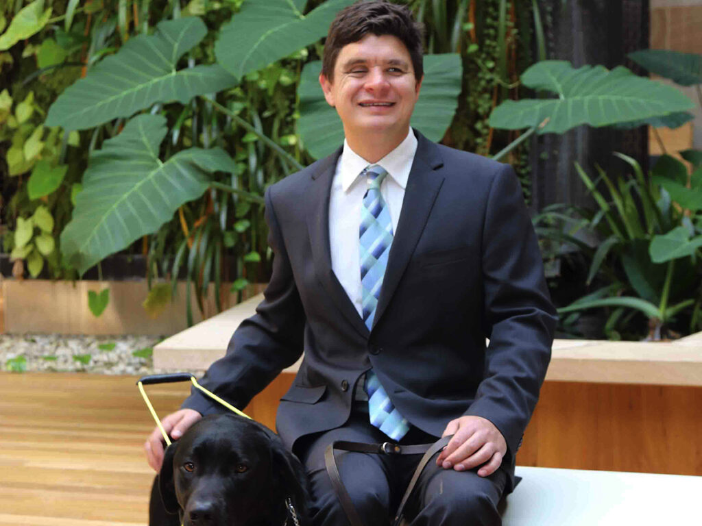 Professor Paul Harper sitting outside in grey suit & blue tie with guide dog