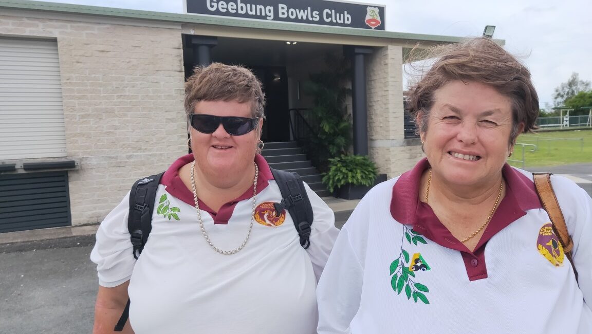 Vanessa & Helen smiling, standing side by side wearing their bowling outfits in-front of a building with a sign reading Welcome to Geebung Bowls Club